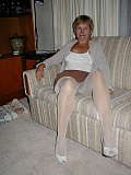 Mature amateur blonde stands confidently in the living room wearing white pantyhose and a white camisole, lifting her skirt to tease her visible bush through the sheer nylon.