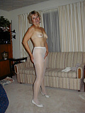 Mature amateur blonde stands confidently in the living room wearing white pantyhose and a white camisole, lifting her skirt to tease her visible bush through the sheer nylon.